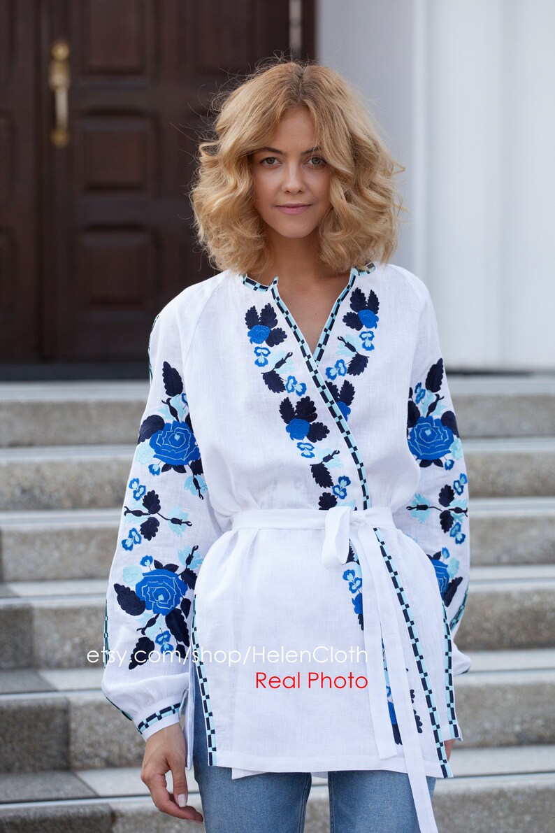 May include: White linen blouse with blue and black floral embroidery. The blouse has a V-neckline and a tie waist. The model is wearing blue jeans. "etsy.com/shop/HelenCloth Real Photo"