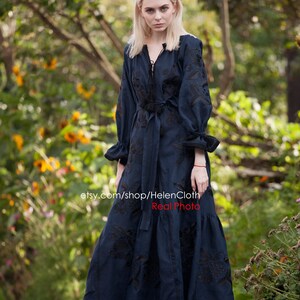 May include: A woman wearing a long, dark blue linen dress with black embroidery. The dress has a V-neckline, long sleeves, and a tie at the waist. The woman is standing in a garden with green trees and yellow flowers in the background. The text "etsy.com/shop/HelenCloth Real Photo" is visible at the bottom of the image.