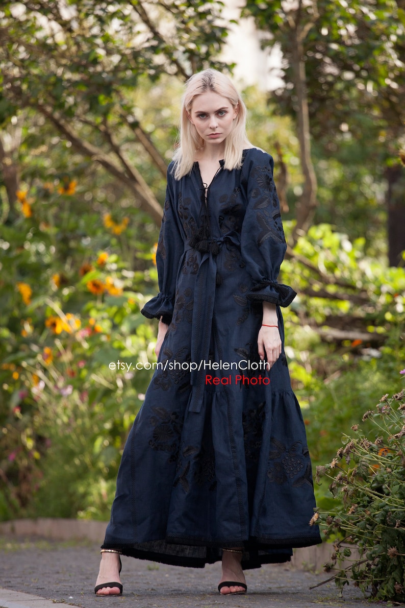 May include: A woman wearing a long, dark blue linen dress with black embroidery. The dress has a V-neckline, long sleeves, and a tie at the waist. The woman is standing in a garden with green trees and yellow flowers in the background. The text "etsy.com/shop/HelenCloth Real Photo" is visible at the bottom of the image.