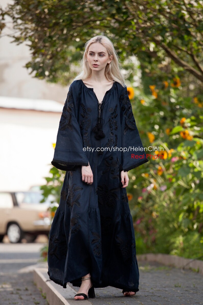 May include: A woman wearing a long, dark blue linen dress with black embroidery. The dress has a V-neckline and long sleeves. The woman is standing on a paved path with a green background. The text "etsy.com/shop/HelenCloth Real Photo" is visible on the dress.