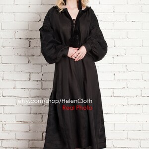 Pu&ograve; includere: Un abito in lino nero con maniche lunghe e scollo arricciato. L'abito ha un laccio al collo e un dettaglio a nappina. La modella indossa l'abito con sandali neri con tacco. Il testo "etsy.com/shop/HelenCloth Real Photo" &egrave; visibile nella parte inferiore dell'immagine.