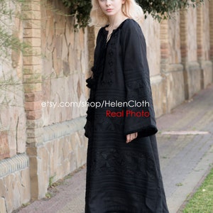 May include: A black linen dress with embroidered geometric patterns. The dress has long sleeves and a loose fit. The model is wearing white sneakers.