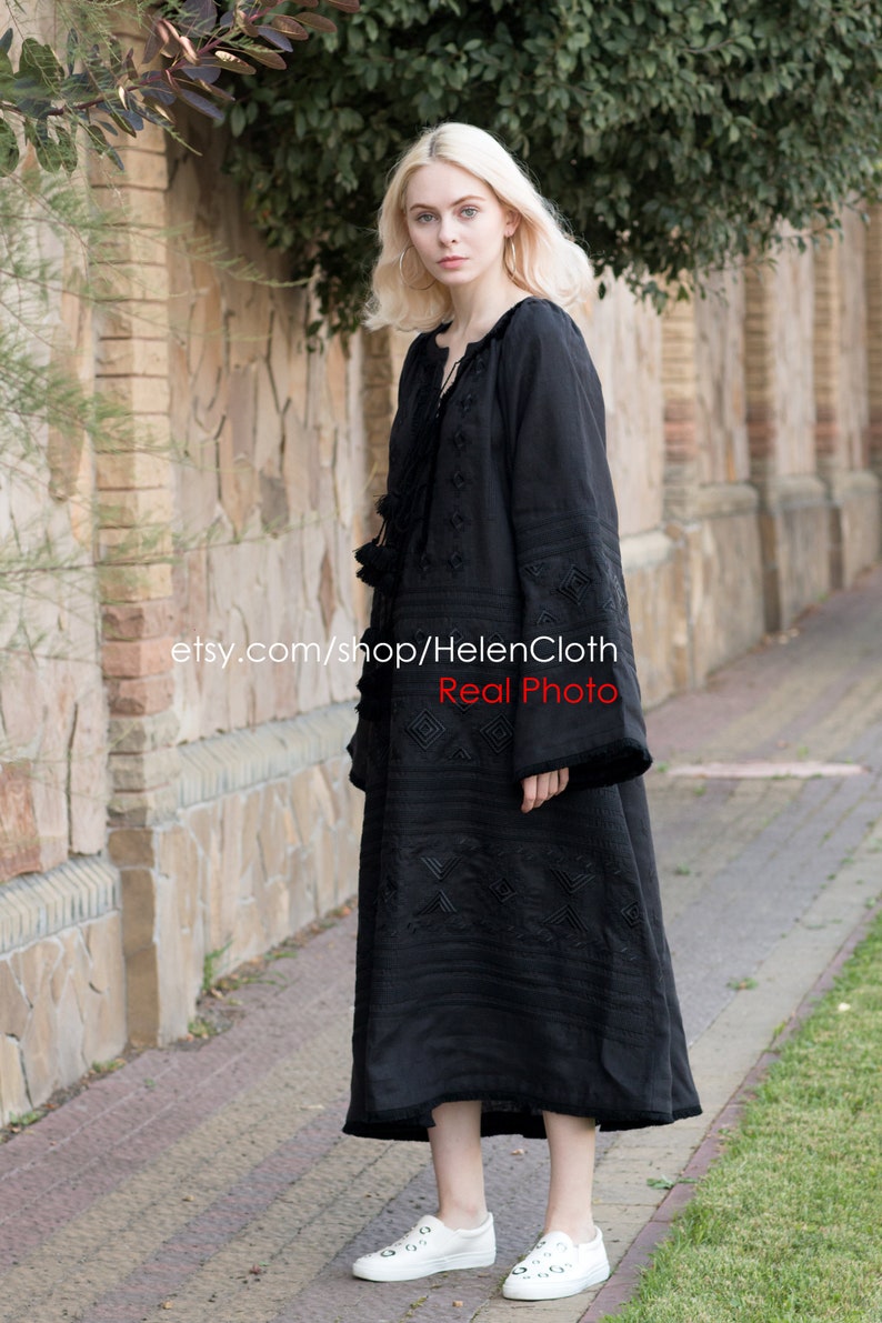 May include: A black linen dress with embroidered geometric patterns. The dress has long sleeves and a loose fit. The model is wearing white sneakers.