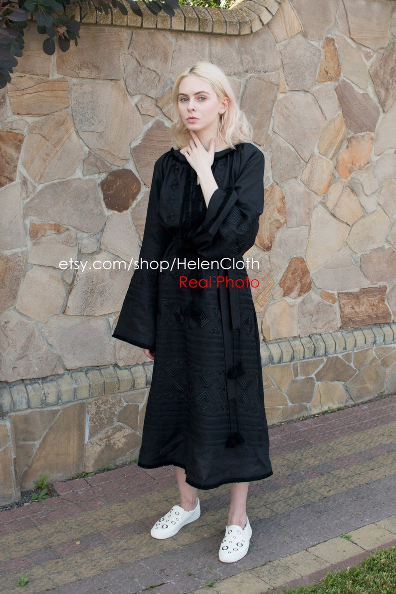 May include: A black embroidered dress with long sleeves and a gathered neckline. The dress has a geometric pattern and is worn by a person standing in front of a stone wall. The text "etsy.com/shop/HelenCloth Real Photo" is visible on the dress.