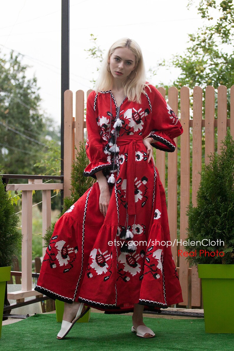 May include: A red embroidered maxi dress with white and black floral patterns. The dress has a long sleeve, a V-neck, and a belt. The model is wearing white sandals. The dress is made of linen and is perfect for summer.