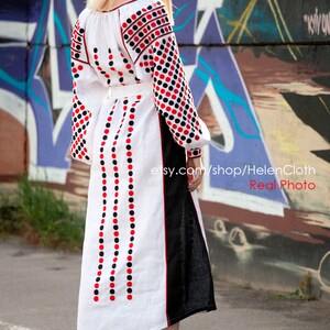 May include: A white linen dress with red and black polka dot embroidery. The dress has a long, flowing skirt and a fitted bodice. The dress is worn by a woman with blonde hair, standing in front of a graffiti wall.