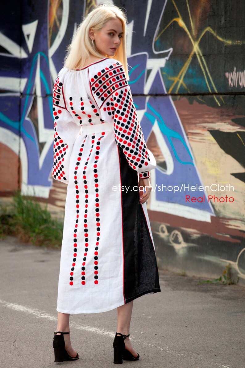 May include: A white linen dress with red and black polka dot embroidery. The dress has a long, flowing skirt and a fitted bodice. The dress is worn by a woman with blonde hair, standing in front of a graffiti wall.