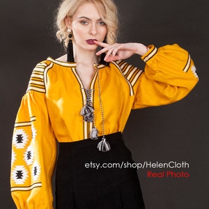 May include: A yellow embroidered blouse with black and white geometric patterns. The blouse has long sleeves and a round neckline. The model is wearing a black skirt and has long blonde hair. The text "etsy.com/shop/HelenCloth Real Photo" is visible at the bottom of the image.