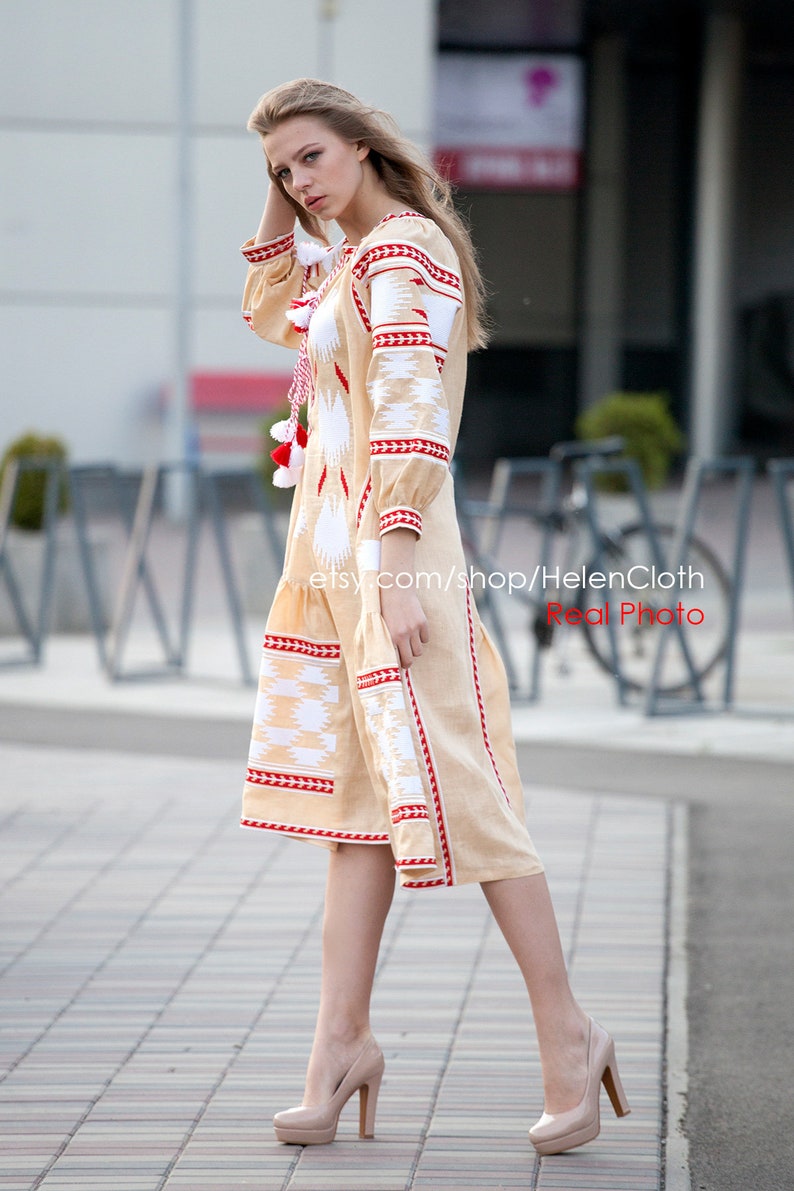 May include: A woman wearing a beige linen dress with white and red embroidery. The dress has a loose fit and a round neckline. The woman is wearing beige high heels.