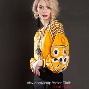 May include: A woman wearing a yellow embroidered blouse with black and white geometric patterns and a black skirt. The blouse has long sleeves and a round neckline. The woman is standing in front of a gray background. etsy.com/shop/HelenCloth Real Photo