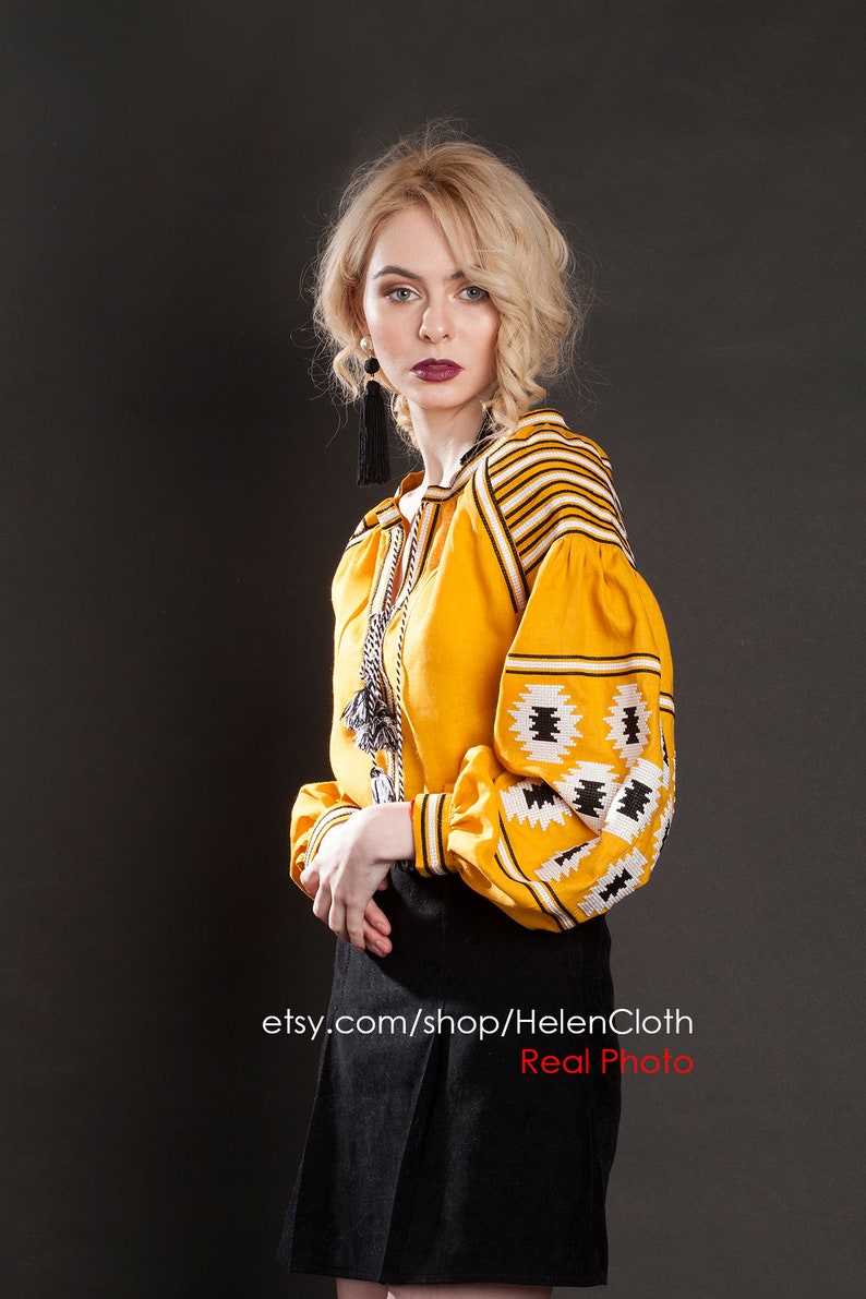May include: A woman wearing a yellow embroidered blouse with black and white geometric patterns and a black skirt. The blouse has long sleeves and a round neckline. The woman is standing in front of a gray background. etsy.com/shop/HelenCloth Real Photo