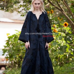 May include: A woman wearing a long, dark blue embroidered dress with black embroidery. The dress has a V-neckline and long sleeves. The woman is standing on a paved path with green foliage in the background. The text "etsy.com/shop/HelenCloth Real Photo" is visible on the dress.
