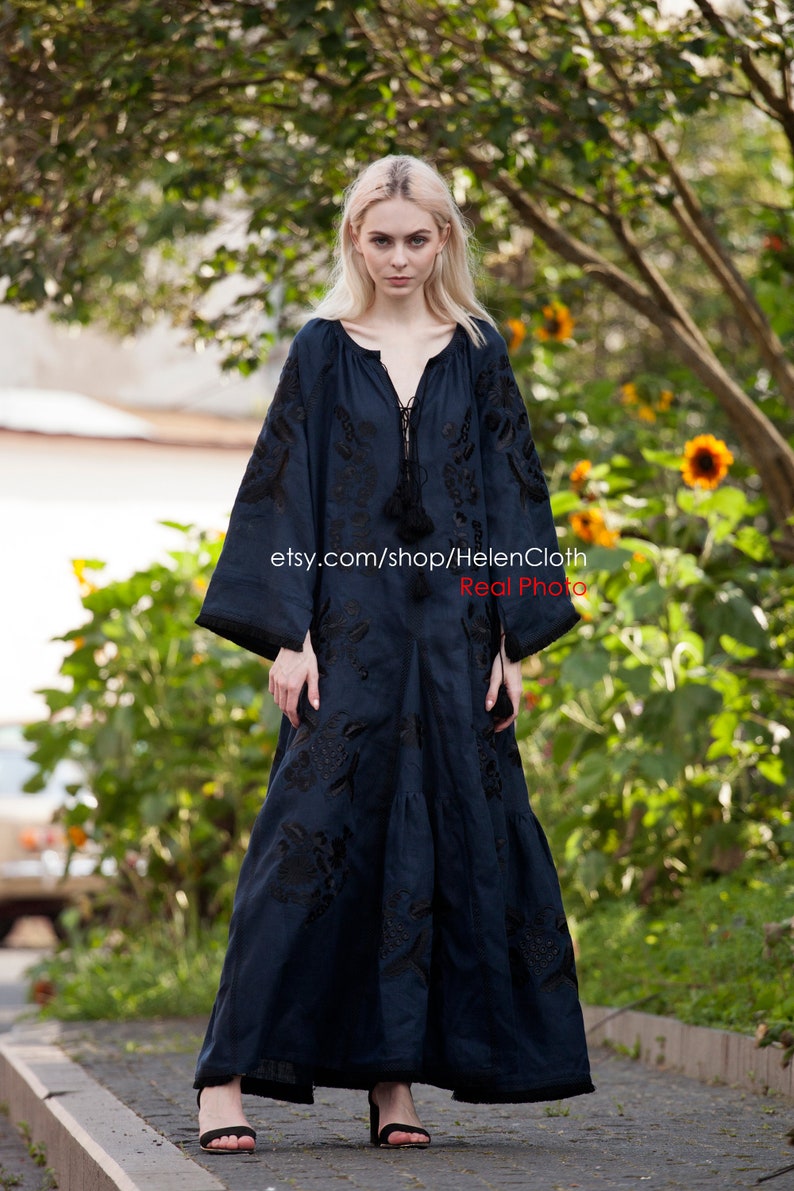 May include: A woman wearing a long, dark blue embroidered dress with black embroidery. The dress has a V-neckline and long sleeves. The woman is standing on a paved path with green foliage in the background. The text "etsy.com/shop/HelenCloth Real Photo" is visible on the dress.