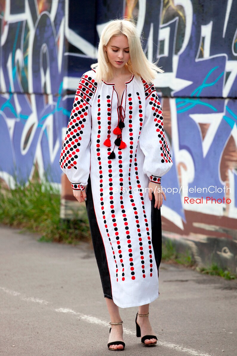 May include: A white embroidered dress with red and black polka dots. The dress has long sleeves and a round neckline. The dress is worn by a person standing in front of a graffiti wall.