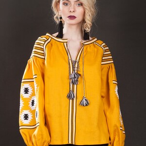May include: A yellow linen blouse with black and white embroidery. The blouse has a V-neck and long sleeves with a tassel detail at the neckline. The model is wearing a black skirt. etsy.com/shop/HelenCloth Real Photo