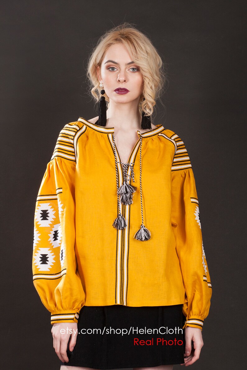 May include: A yellow linen blouse with black and white embroidery. The blouse has a V-neck and long sleeves with a tassel detail at the neckline. The model is wearing a black skirt. etsy.com/shop/HelenCloth Real Photo