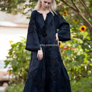 May include: A long, dark blue maxi dress with black embroidery. The dress has a round neckline and long sleeves. The model is wearing the dress with black heeled sandals. The dress is photographed outdoors in front of a green background.
