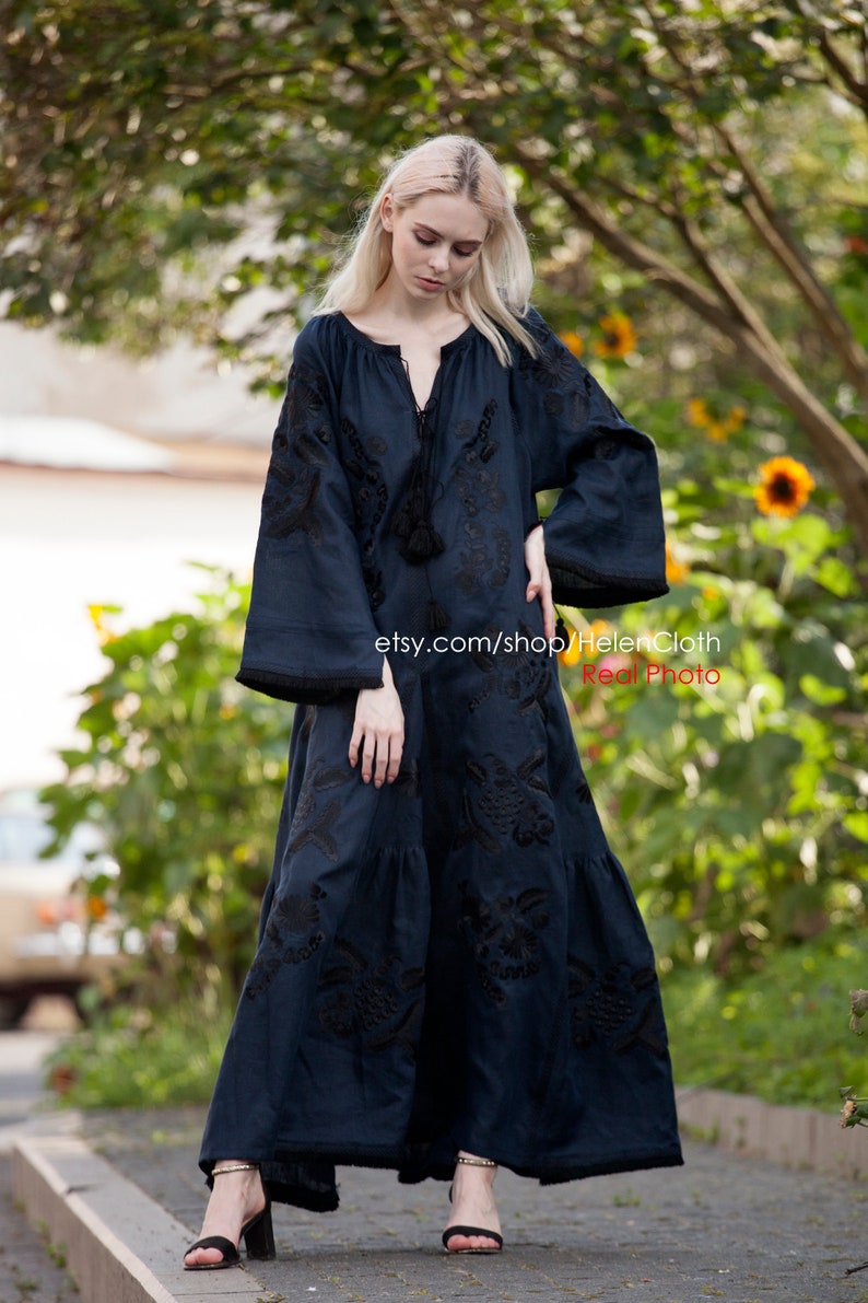 May include: A long, dark blue maxi dress with black embroidery. The dress has a round neckline and long sleeves. The model is wearing the dress with black heeled sandals. The dress is photographed outdoors in front of a green background.