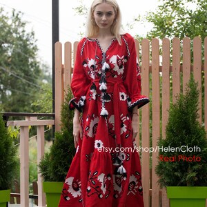 May include: A woman wearing a red embroidered maxi dress with white and black floral patterns. The dress has a long sleeve and a cinched waist. The woman is standing in front of a wooden fence and green plants.