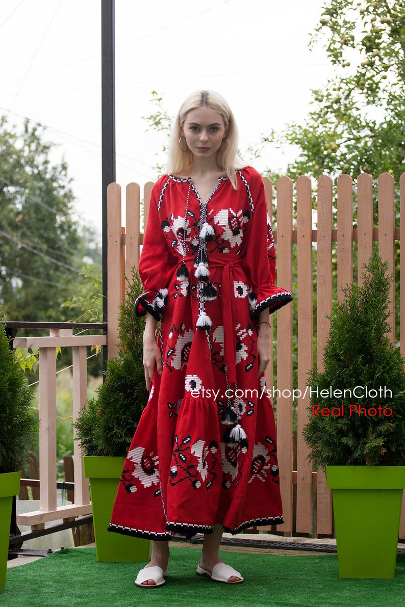 May include: A woman wearing a red embroidered maxi dress with white and black floral patterns. The dress has a long sleeve and a cinched waist. The woman is standing in front of a wooden fence and green plants.