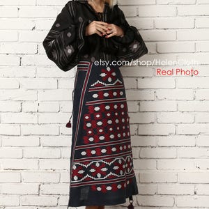 May include: A woman wearing a black embroidered blouse with long sleeves and a dark blue skirt with red and white geometric patterns. The skirt has a tassel detail. She is standing in front of a white brick wall.