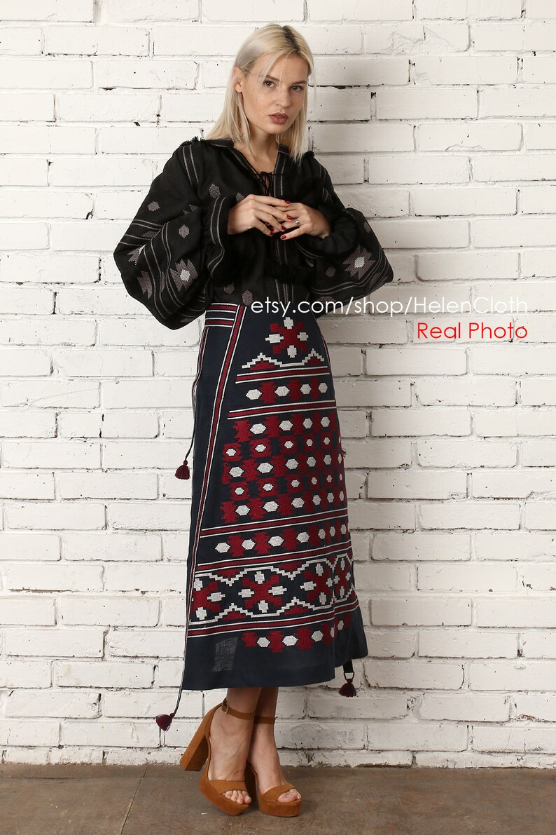 May include: A woman wearing a black embroidered blouse with long sleeves and a dark blue skirt with red and white geometric patterns. The skirt has a tassel detail. She is standing in front of a white brick wall.