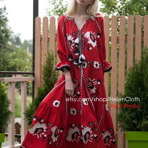 May include: A red maxi dress with white and black embroidery. The dress has a V-neckline and long sleeves. The model is wearing white sandals. The dress is made of linen and is perfect for summer.