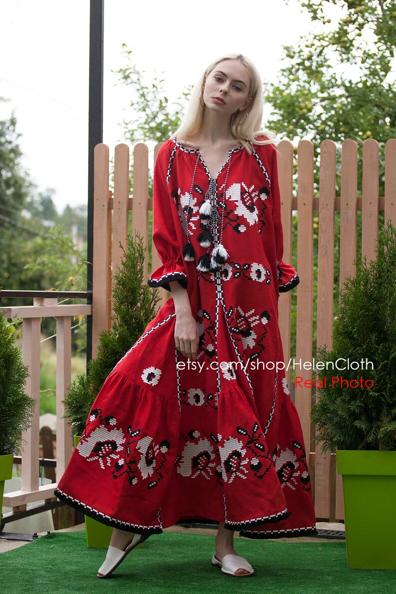 May include: A red maxi dress with white and black embroidery. The dress has a V-neckline and long sleeves. The model is wearing white sandals. The dress is made of linen and is perfect for summer.