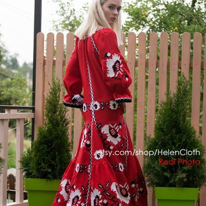 May include: A red linen dress with white and black embroidery. The dress has a long skirt and a fitted bodice. The model is wearing the dress outdoors in front of a wooden fence.