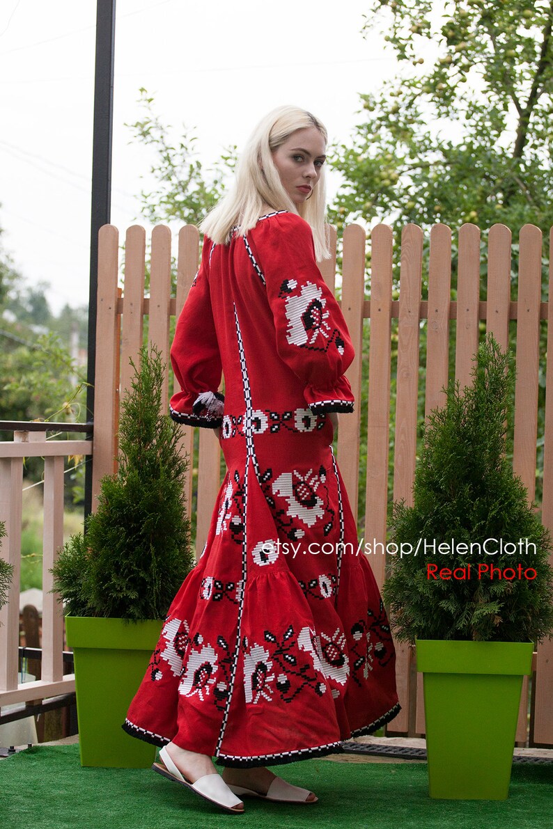May include: A red linen dress with white and black embroidery. The dress has a long skirt and a fitted bodice. The model is wearing the dress outdoors in front of a wooden fence.