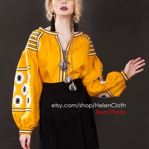 May include: A yellow embroidered linen blouse with black and white geometric patterns. The blouse has long sleeves with a gathered cuff and a V-neckline. The model is wearing a black skirt and black tassel earrings. The image includes the text "etsy.com/shop/HelenCloth Real Photo".