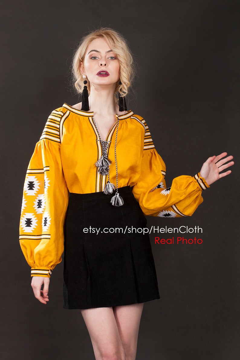 May include: A yellow embroidered linen blouse with black and white geometric patterns. The blouse has long sleeves with a gathered cuff and a V-neckline. The model is wearing a black skirt and black tassel earrings. The image includes the text "etsy.com/shop/HelenCloth Real Photo".