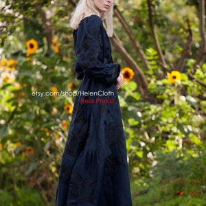 May include: A woman wearing a dark blue maxi dress with black embroidery. The dress has a ruffled skirt and long sleeves. She is standing on a paved path with a green background of trees and yellow flowers.