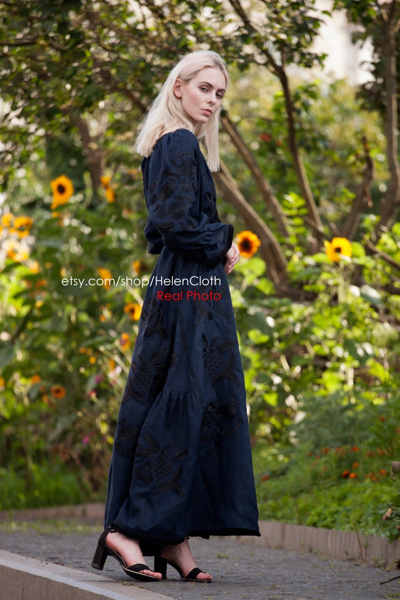 May include: A woman wearing a dark blue maxi dress with black embroidery. The dress has a ruffled skirt and long sleeves. She is standing on a paved path with a green background of trees and yellow flowers.