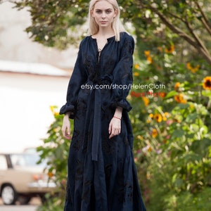 May include: A woman wearing a long, dark blue linen dress with black embroidery. The dress has a V-neckline and a tie at the waist. The woman is standing on a sidewalk in front of a green bush.