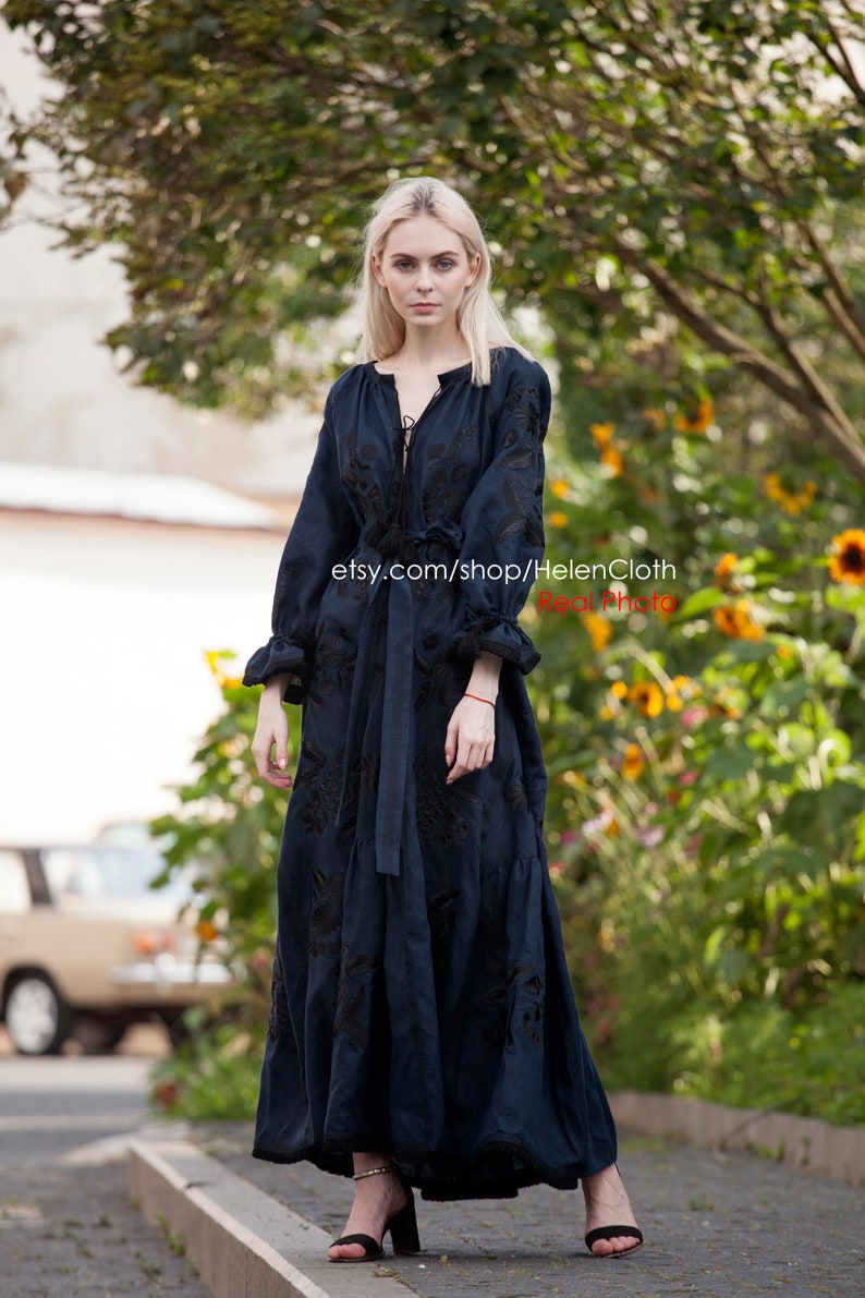 May include: A woman wearing a long, dark blue linen dress with black embroidery. The dress has a V-neckline and a tie at the waist. The woman is standing on a sidewalk in front of a green bush.