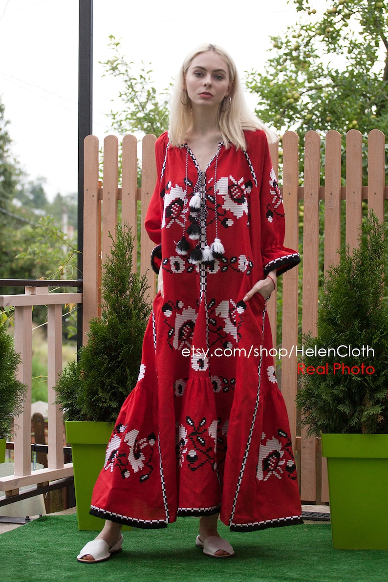 May include: A red linen dress with white and black embroidery. The dress has a long, flowing skirt and a fitted bodice. The dress is decorated with tassels and has a V-neckline. The dress is worn by a woman with blonde hair. The image is taken outdoors and shows a wooden fence and green plants in the background. The text "etsy.com/shop/HelenCloth Real Photo" is visible on the dress.