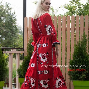 May include: A red embroidered linen dress with white and black floral patterns. The dress has a long skirt and a fitted bodice. The model is wearing white sandals and standing in front of a wooden fence.
