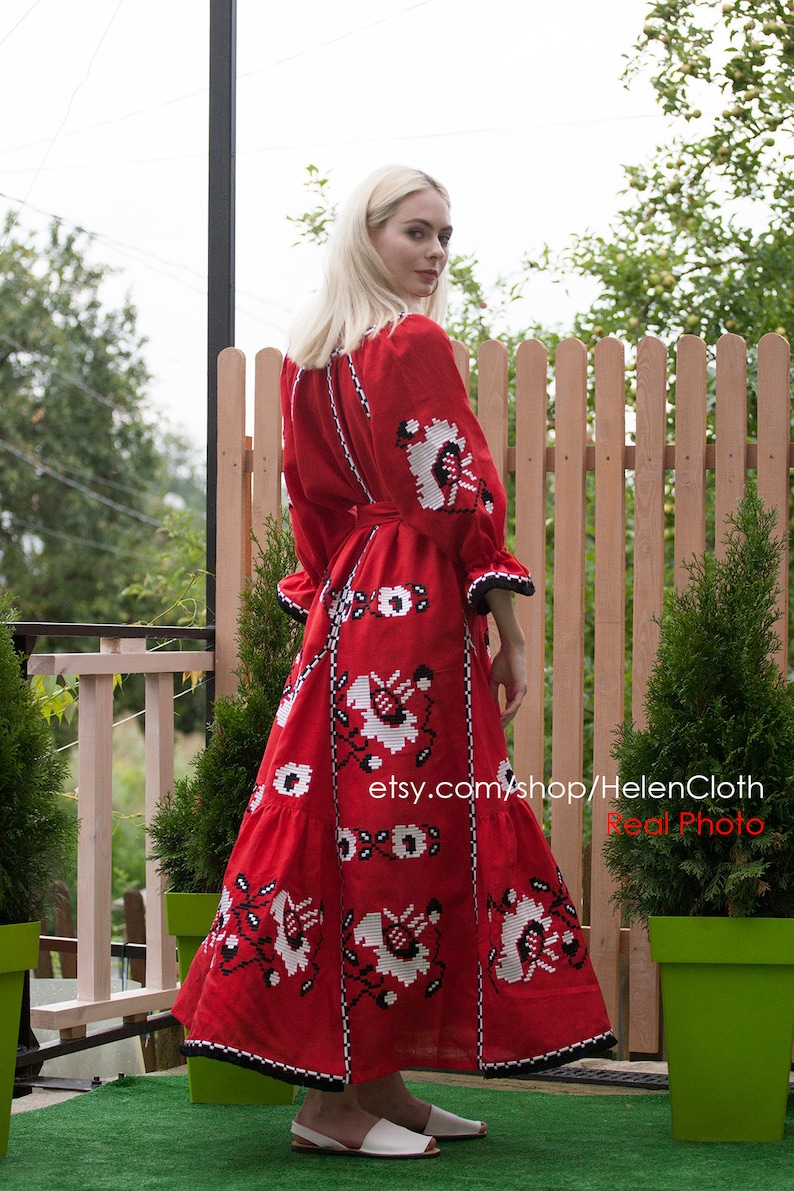 May include: A red embroidered linen dress with white and black floral patterns. The dress has a long skirt and a fitted bodice. The model is wearing white sandals and standing in front of a wooden fence.