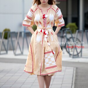 May include: A woman wearing a beige linen dress with white and red embroidery. The dress has a fitted bodice and a flowing skirt. The woman is standing on a sidewalk with a blurred background.
