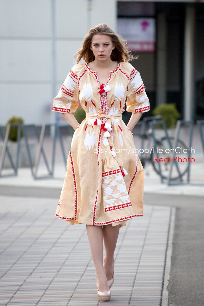 May include: A woman wearing a beige linen dress with white and red embroidery. The dress has a fitted bodice and a flowing skirt. The woman is standing on a sidewalk with a blurred background.