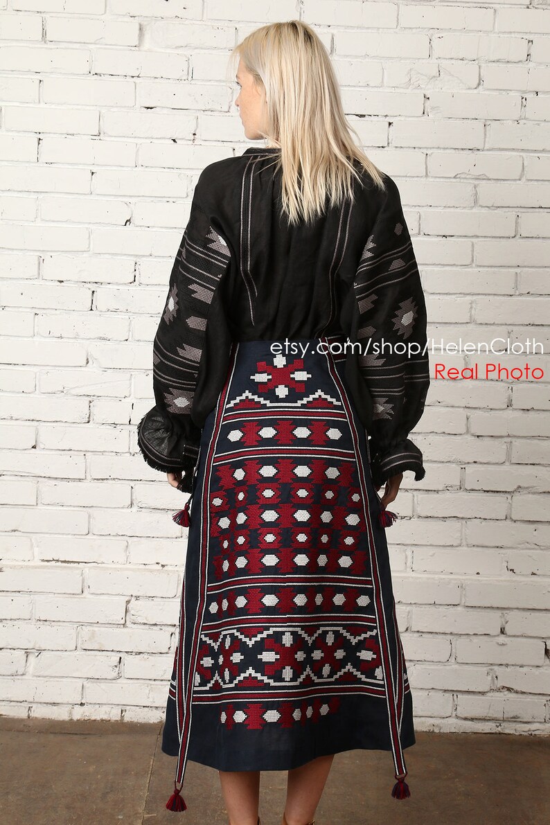May include: A black linen blouse with embroidered geometric patterns and a long, dark blue skirt with red and white embroidered geometric patterns. The skirt has tassels on the bottom.