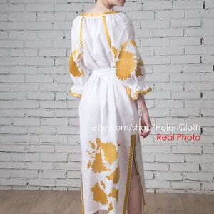 May include: A white linen dress with gold embroidery and a side slit. The dress has a belt and tassels on the bottom. The dress is photographed on a model against a white brick wall. The text "etsy.com/shop/HelenCloth Real Photo" is visible at the bottom of the image.