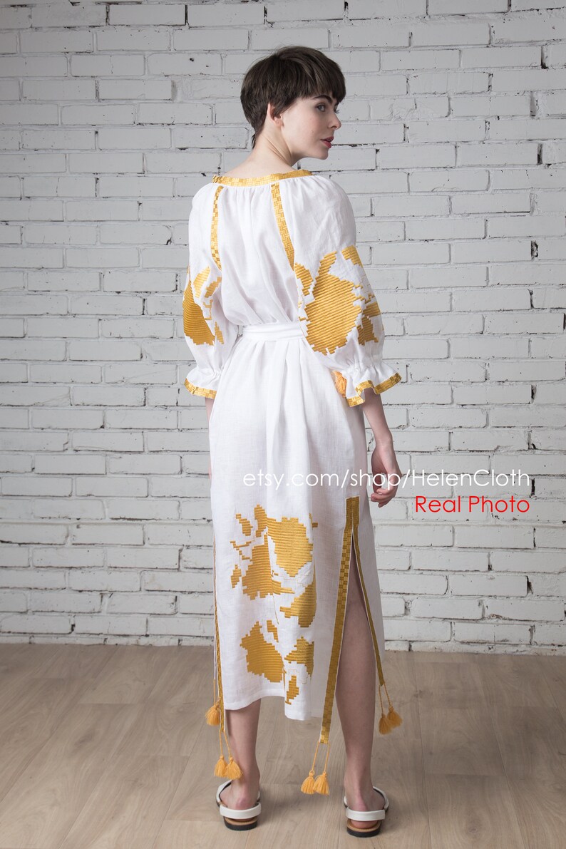 May include: A white linen dress with gold embroidery and a side slit. The dress has a belt and tassels on the bottom. The dress is photographed on a model against a white brick wall. The text "etsy.com/shop/HelenCloth Real Photo" is visible at the bottom of the image.