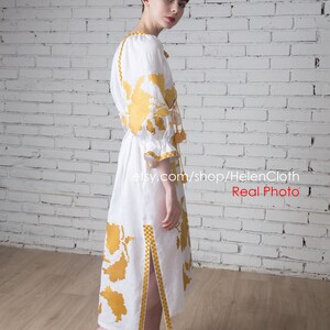 May include: A white linen dress with yellow embroidered floral patterns. The dress has a long, flowing skirt and a fitted bodice. The dress is cinched at the waist with a yellow ribbon. The dress has a side slit and yellow tassels on the sleeves and hem.