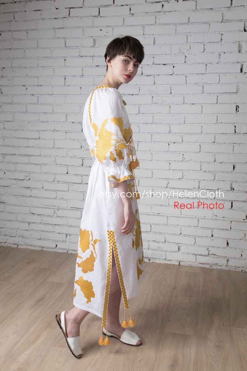 May include: A white linen dress with yellow embroidered floral patterns. The dress has a long, flowing skirt and a fitted bodice. The dress is cinched at the waist with a yellow ribbon. The dress has a side slit and yellow tassels on the sleeves and hem.