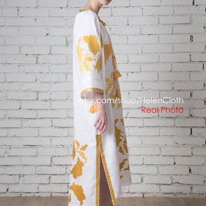 May include: White linen dress with yellow embroidered floral pattern and tassels. The dress has a side slit and is worn by a person with short dark hair. The background is a white brick wall.