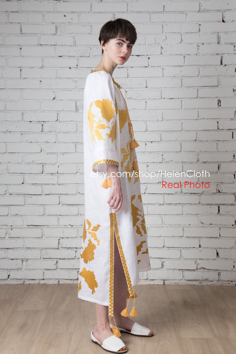 May include: White linen dress with yellow embroidered floral pattern and tassels. The dress has a side slit and is worn by a person with short dark hair. The background is a white brick wall.