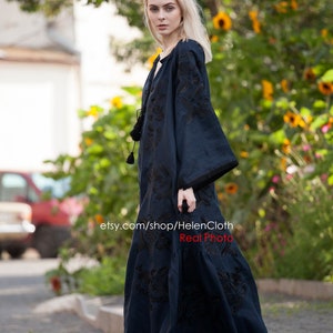 May include: A woman wearing a long, dark blue embroidered dress with black embroidery. The dress has long sleeves and a loose fit. The woman is standing on a sidewalk in front of a green bush.