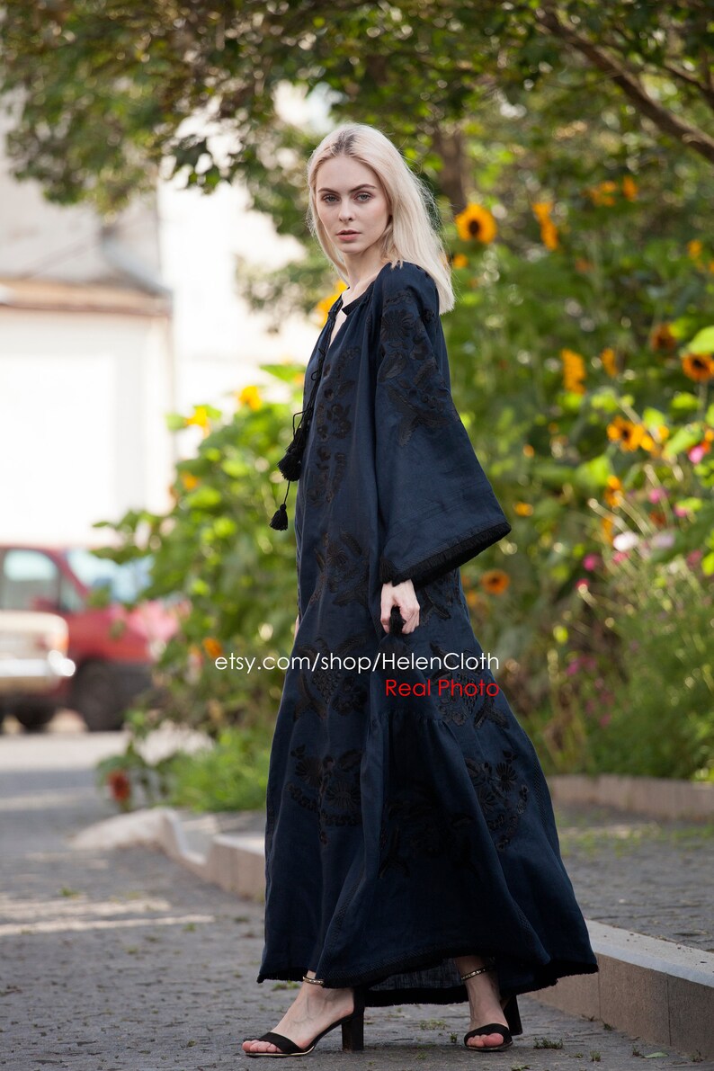 May include: A woman wearing a long, dark blue embroidered dress with black embroidery. The dress has long sleeves and a loose fit. The woman is standing on a sidewalk in front of a green bush.